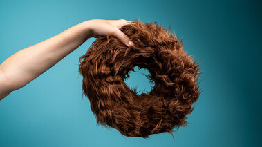 Hand hält einen torusförmigen Gegenstand aus braunen Fasern vor blauem Hintergrund.